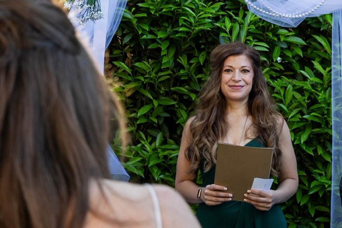 A woman in a green dress holds a clipboard and smiles as a woman wearing a white bridal dress approaches.